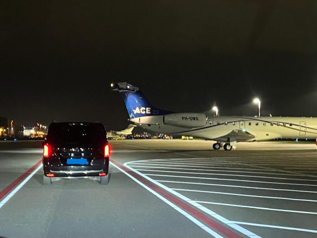 Black van driving on the tarmac toward a parked airplane at night.